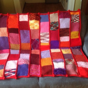 Könnte beinhalten: Eine bunte gehäkelte Decke mit Patchwork-Muster. Die Decke besteht aus Quadraten in verschiedenen Farben, darunter Rot, Orange, Lila, Rosa und Braun. Die Decke liegt auf einem grauen Sofa.