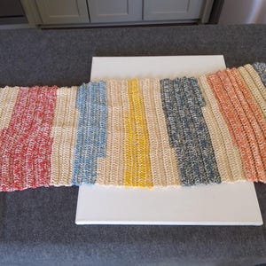 May include: A colorful, hand-crocheted table runner. The runner features vertical stripes in red, blue, yellow, cream, and orange. The textured yarn creates a unique, handcrafted look. This decorative piece adds a pop of color to any table setting.