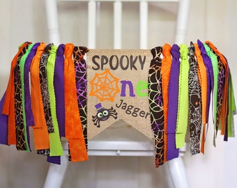 Op de afbeelding: Een banner van jute met de woorden "Spooky One" en een spinnenafbeelding. De banner is versierd met paarse, groene, oranje en zwarte stoffen streamers.