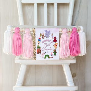 May include: A high chair banner featuring a rectangular sign with the text Audrey's First Chapter surrounded by various storybook character illustrations. The banner includes wooden beads and alternating white, light pink, and dark pink yarn tassels.