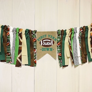 May include: A burlap banner with the text "First Year Owen -Down- " and a football graphic. The banner is decorated with green, brown, and white fabric strips.