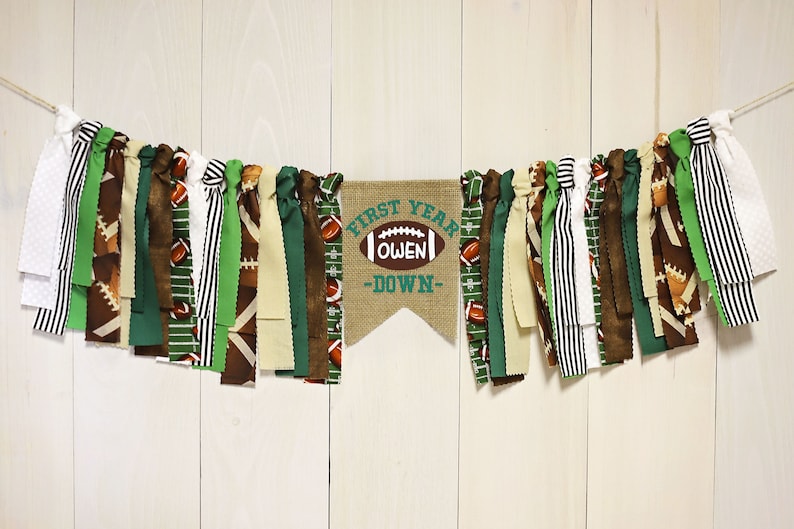 May include: A burlap banner with the text "First Year Owen -Down- " and a football graphic. The banner is decorated with green, brown, and white fabric strips.