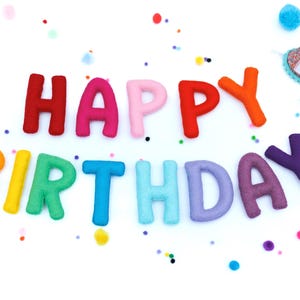 May include: A set of felt letters spelling out HAPPY BIRTHDAY in various colors including red, pink, orange, yellow, green, blue, and purple, accompanied by small colorful pom-poms and a small party hat with the number one on it.