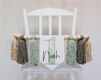 May include: A white high chair with a green and brown tassel garland and a wooden banner that says "1 Noah".