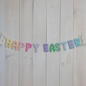 May include: A pastel colored felt banner with the words "Happy Easter" hanging on a white wood background. The banner is decorated with two white felt bunnies.