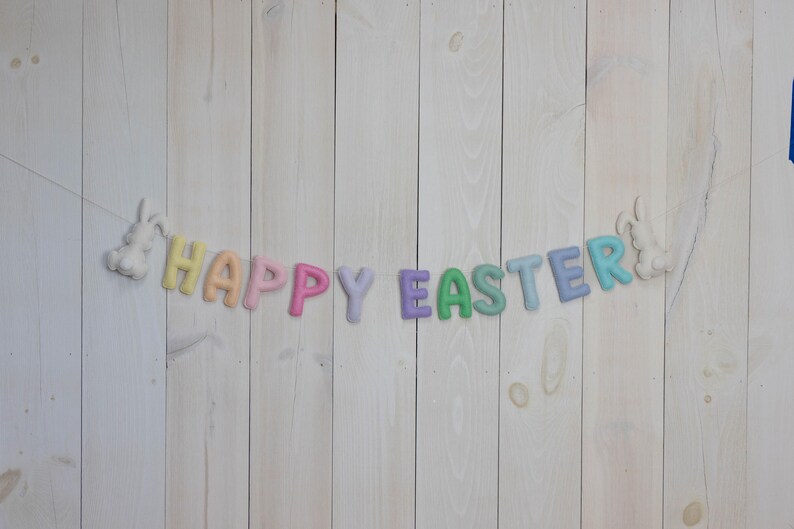 May include: A pastel colored felt banner with the words "Happy Easter" hanging on a white wood background. The banner is decorated with two white felt bunnies.