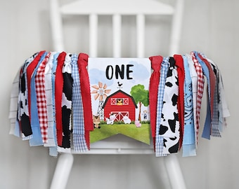 May include: A red, white, and blue fabric banner with the word "ONE" on it. The banner has a farm theme with a red barn, a windmill, and farm animals. The banner is decorated with red, white, and blue fabric strips.