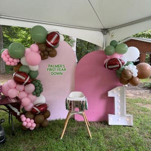 May include: A pink and green balloon arch with football balloons and a sign that says "Palmer's First Year Down". There is a white light-up number one and a high chair with a green and brown fringe.