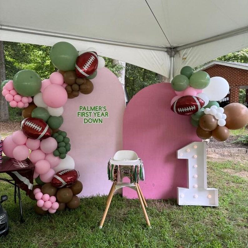May include: A pink and green balloon arch with football balloons and a sign that says "Palmer's First Year Down". There is a white light-up number one and a high chair with a green and brown fringe.