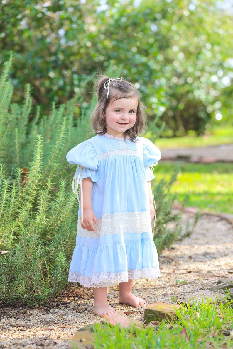 Puede incluir: Una ni&ntilde;a peque&ntilde;a con un vestido azul claro con ribete de encaje blanco y un lazo blanco en el pelo. Est&aacute; de pie en un jard&iacute;n con plantas verdes y piedras marrones.