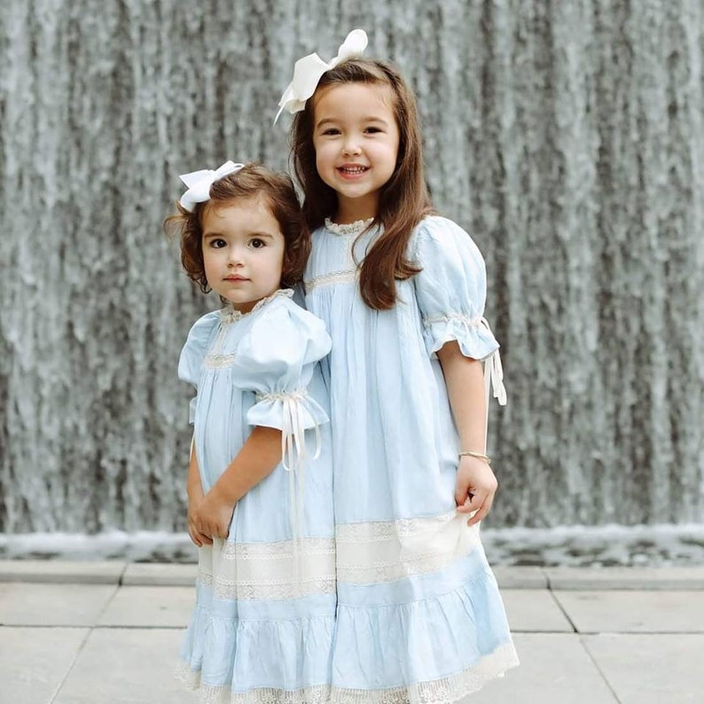 Puede incluir: Dos ni&ntilde;as peque&ntilde;as con vestidos a juego de color azul claro con ribetes de encaje blanco y lazos blancos en el pelo. Las ni&ntilde;as est&aacute;n de pie frente a una fuente de agua.