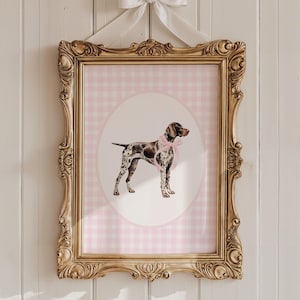 May include: A framed print featuring a German Shorthaired Pointer wearing a pink bow, set against a pink gingham background. The ornate gold frame is topped with a white bow. A decorative piece for a nursery or pet lover.