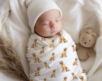 May include: A newborn baby wearing a white knit hat and a swaddle blanket with a pattern of golden retriever dogs. The baby is sleeping peacefully and is surrounded by a white blanket.