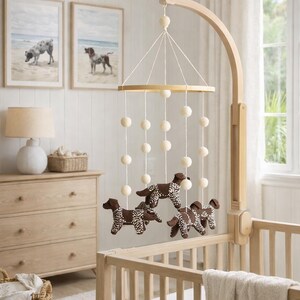 Op de afbeelding: Een houten babybedje met een mobiel met bruine hondenfiguren met witte stippen. De mobiel hangt aan een houten arm. Twee ingelijste hondenportretten hangen aan de muur. De kamer heeft een licht, neutraal kleurenschema.