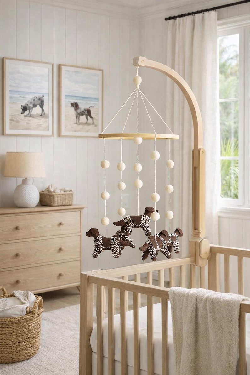 Op de afbeelding: Een houten babybedje met een mobiel met bruine hondenfiguren met witte stippen. De mobiel hangt aan een houten arm. Twee ingelijste hondenportretten hangen aan de muur. De kamer heeft een licht, neutraal kleurenschema.
