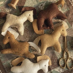 May include: A collection of four felt horse ornaments in shades of cream, tan, and dark brown, each featuring a contrasting mane and tail. A wooden spool and a pair of gold-toned metal scissors are positioned alongside the plush horse figures.