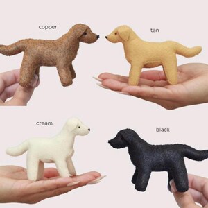 Op de afbeelding: Vier vilten hondenbeeldjes in koper, bruin, cr&egrave;me en zwart. Elke hond staat, met de kleurnaam erboven. De honden zijn klein en lijken handgemaakt.
