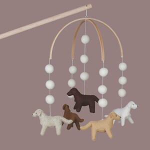 Op de afbeelding: Een baby mobile met vilten honden in verschillende kleuren, hangend onder witte pompons en een houten boog. Een charmant decoratief element voor de kinderkamer.