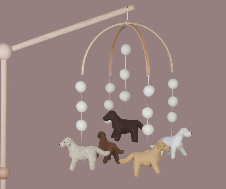 Op de afbeelding: Een baby mobile met vilten honden in verschillende kleuren, hangend onder witte pompons en een houten boog. Een charmant decoratief element voor de kinderkamer.