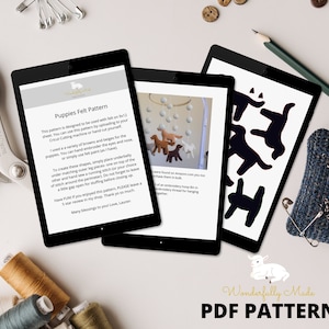 Op de afbeelding: Drie tablets tonen een digitaal PDF-patroon voor vilten puppy's. Het patroon bevat instructies voor handborduurwerk of viltverfdetails. De afbeelding toont ook knutselgereedschap zoals schaar, draad en een speldenkussen.