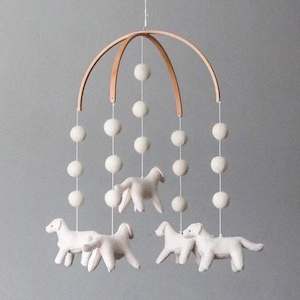 May include: A white felt baby mobile with a wooden frame and white felt dogs hanging from it. The mobile is decorated with white pom-poms.