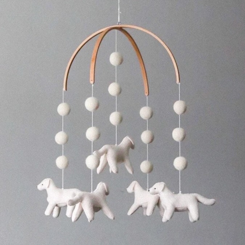 May include: A white felt baby mobile with a wooden frame and white felt dogs hanging from it. The mobile is decorated with white pom-poms.
