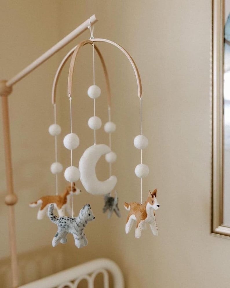 May include: A wooden baby mobile with a crescent moon, white pom poms, and felt animal figures. The animals include a grey and white cat, a brown and white dog, and a brown and white dog with a black nose.