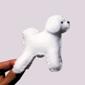 Puede incluir: Una pequeña figura de perro de fieltro blanco con ojos de cuentas negras y una nariz negra. El perro está de perfil, mostrando sus rasgos suaves y redondeados. El juguete está sostenido por una mano.