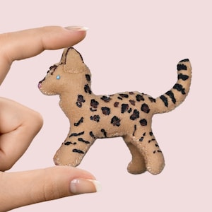 May include: A small, brown and black felt toy cat with a spotted pattern. The cat is standing on its four paws and has a blue eye.