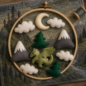 May include: A wooden embroidery hoop containing felt shapes including a green dragon, two grey mountains with white peaks, two dark green pine trees, a cream crescent moon, and four white clouds, all arranged against a fabric background.