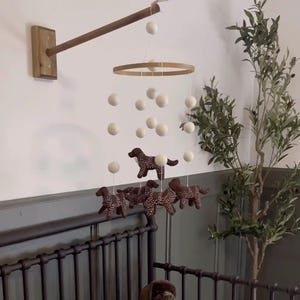 Op de afbeelding: Een baby mobile met bruine hondenfiguren met witte stippen, hangend onder witte bollen en een houten ring. De mobile is bevestigd aan een houten arm en gemonteerd aan een muur, boven een donker metalen ledikant.