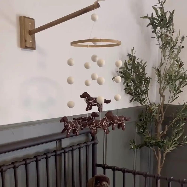 Op de afbeelding: Een baby mobile met bruine hondenfiguren met witte stippen, hangend onder witte bollen en een houten ring. De mobile is bevestigd aan een houten arm en gemonteerd aan een muur, boven een donker metalen ledikant.