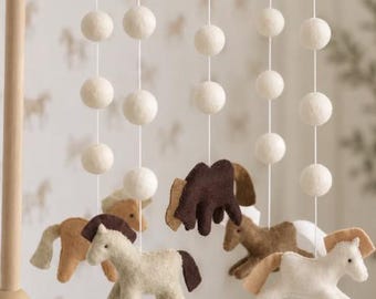 Könnte beinhalten: Ein Kinderzimmer-Mobile mit Filz-Pferden in Weiß-, Beige- und Dunkelbrauntönen, die an weißen Schnüren an einem Holzrahmen hängen. Das Mobile enthält mehrere weiße Filzkugeln, die vertikal über jeder Pferdefigur angeordnet sind.