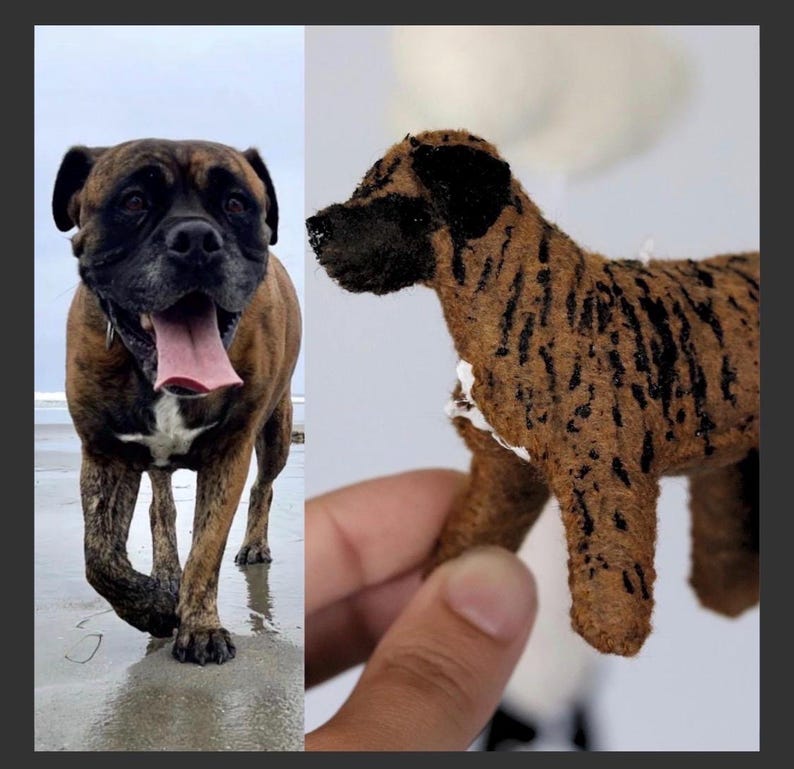 Op de afbeelding: Een gestroomde Boxerhond met een zwarte snuit en een roze tong, op het strand. Daarnaast een klein, handgemaakt hondenbeeldje met bruine en zwarte markeringen.