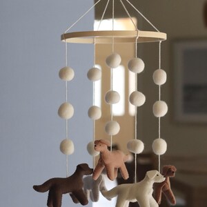 K&ouml;nnte beinhalten: Ein h&auml;ngendes Mobile mit einem runden Holzrahmen und daran befestigten wei&szlig;en Filzkugeln an Schn&uuml;ren. Vier Filzhunde in den Farben Dunkelbraun, Hellbraun, Creme und Beige h&auml;ngen an den Schn&uuml;ren und bilden eine geschichtete Anordnung.