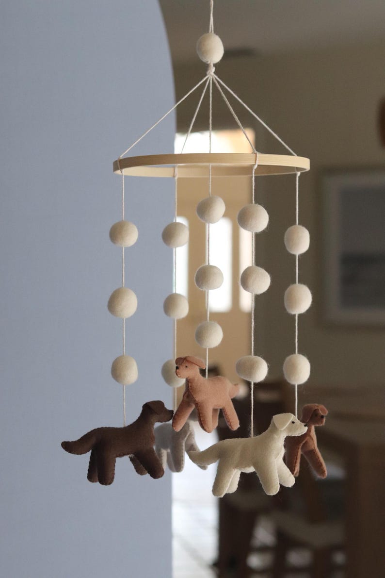 K&ouml;nnte beinhalten: Ein h&auml;ngendes Mobile mit einem runden Holzrahmen und daran befestigten wei&szlig;en Filzkugeln an Schn&uuml;ren. Vier Filzhunde in den Farben Dunkelbraun, Hellbraun, Creme und Beige h&auml;ngen an den Schn&uuml;ren und bilden eine geschichtete Anordnung.