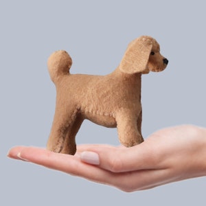 Puede incluir: Una mano sosteniendo un pequeño caniche de juguete de fieltro marrón. El caniche está de pie sobre sus cuatro patas y tiene una cola esponjosa.