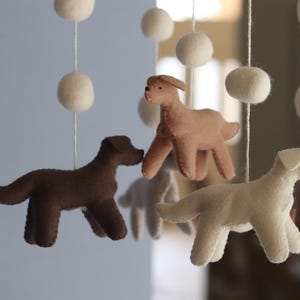 K&ouml;nnte beinhalten: Ein h&auml;ngendes Mobile mit mehreren Filzhundefiguren in Braun-, Beige- und Cremet&ouml;nen, die an wei&szlig;en Schn&uuml;ren befestigt sind, welche mit runden wei&szlig;en Filzkugeln durchsetzt sind. Die Hunde sind in verschiedenen stehenden Posen dargestellt.