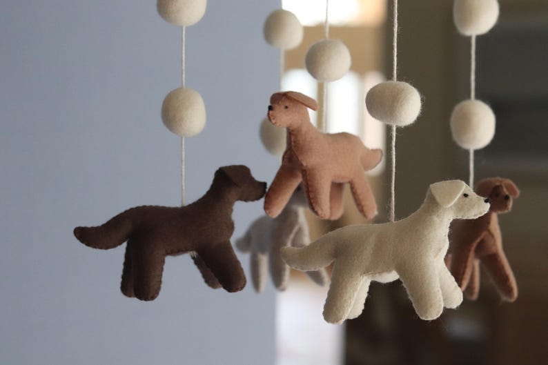 K&ouml;nnte beinhalten: Ein h&auml;ngendes Mobile mit mehreren Filzhundefiguren in Braun-, Beige- und Cremet&ouml;nen, die an wei&szlig;en Schn&uuml;ren befestigt sind, welche mit runden wei&szlig;en Filzkugeln durchsetzt sind. Die Hunde sind in verschiedenen stehenden Posen dargestellt.