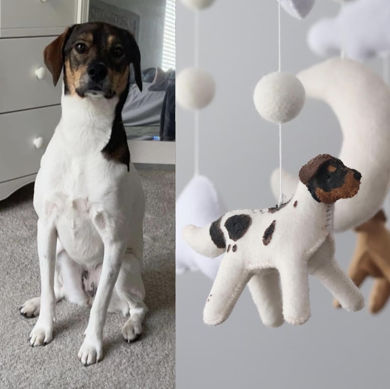 Op de afbeelding: Een wit-bruine hond zit op een grijs tapijt naast een babymobiel. De mobiel heeft een vilten hond met zwarte vlekken, een maan, witte wolken en witte pompons. De hond in de mobiel heeft een vergelijkbaar patroon als de echte hond.