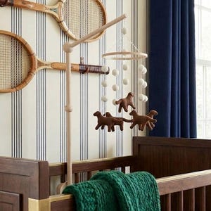 Op de afbeelding: Een houten babybedje met een groene deken, een mobiel met bruine paardenfiguren en witte ballen, en twee vintage tennisrackets aan een gestreepte muur. Donkerblauwe gordijnen zijn zichtbaar op de achtergrond.