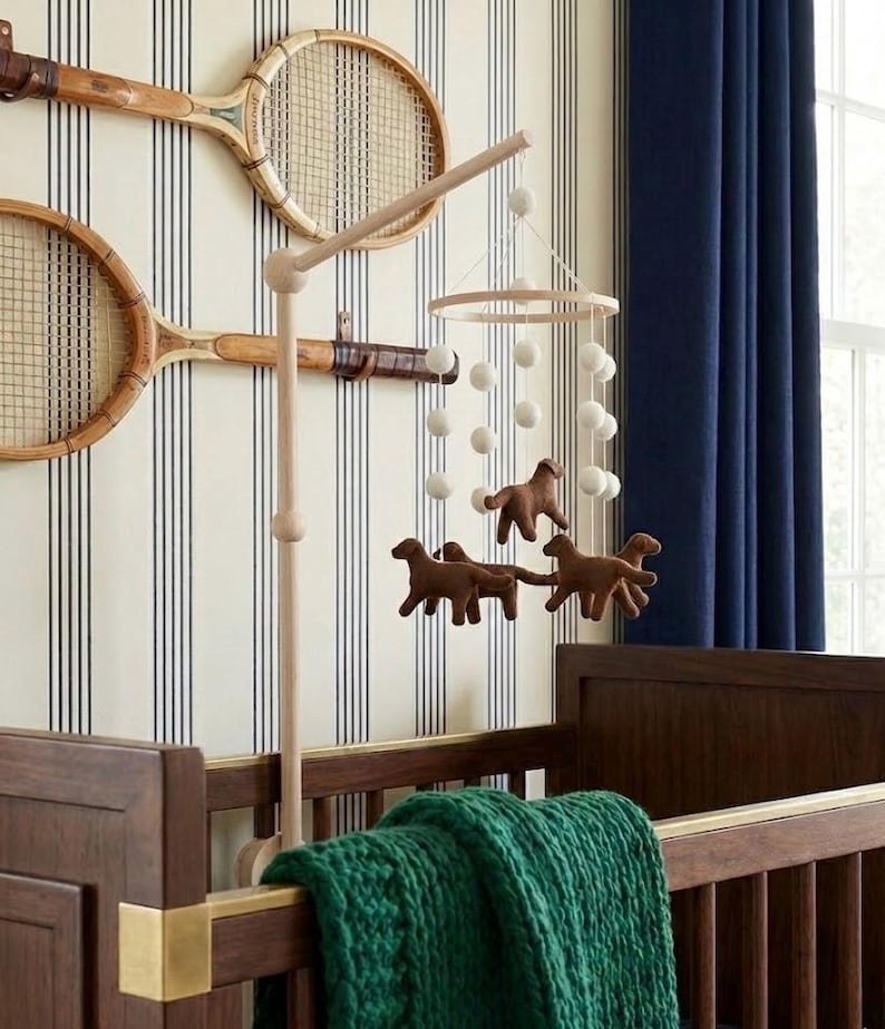 Op de afbeelding: Een houten babybedje met een groene deken, een mobiel met bruine paardenfiguren en witte ballen, en twee vintage tennisrackets aan een gestreepte muur. Donkerblauwe gordijnen zijn zichtbaar op de achtergrond.