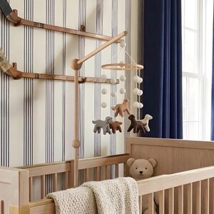 Op de afbeelding: Een houten babybedje met een beige gebreide deken en een teddybeer. Boven het bedje hangt een mobiel met vilten dierenfiguren en witte pompons. De kamer heeft gestreept behang en blauwe gordijnen.