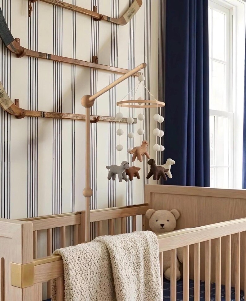 Op de afbeelding: Een houten babybedje met een beige gebreide deken en een teddybeer. Boven het bedje hangt een mobiel met vilten dierenfiguren en witte pompons. De kamer heeft gestreept behang en blauwe gordijnen.