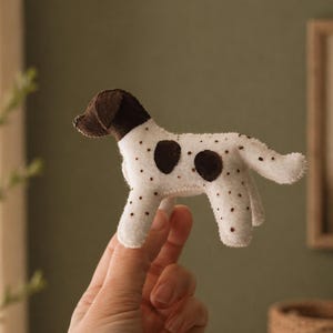 Op de afbeelding: Een handgemaakt hondenknuffel van vilt, wit met bruine vlekken en een bruine kop. Het speelgoed wordt door een hand omhoog gehouden, wat de kleine afmetingen en gedetailleerde afwerking benadrukt. Het is een charmant, zacht en knuffelbaar item.