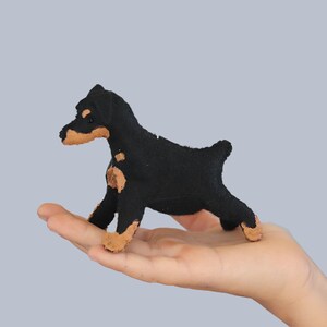 Puede incluir: Un pequeño juguete de peluche de perro negro y marrón. El perro está de pie sobre sus patas traseras con las patas delanteras levantadas.