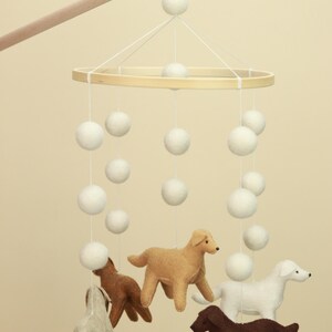 Op de afbeelding: Een baby mobile met een houten ring en witte vilten ballen die aan touwtjes hangen. Daaronder hangen vilten hondenfiguren in verschillende kleuren, waaronder beige, bruin en wit. De mobile is opgehangen aan een houten deuvel.
