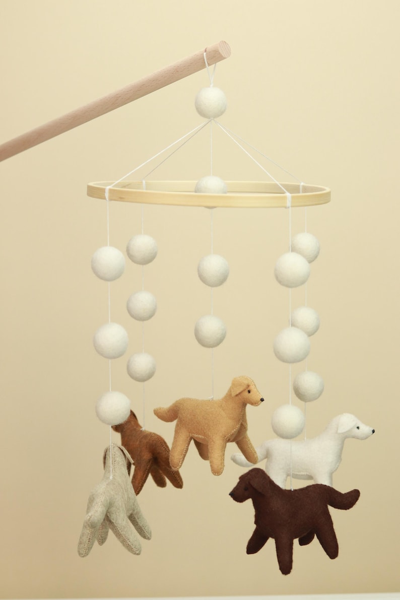 Op de afbeelding: Een baby mobile met een houten ring en witte vilten ballen die aan touwtjes hangen. Daaronder hangen vilten hondenfiguren in verschillende kleuren, waaronder beige, bruin en wit. De mobile is opgehangen aan een houten deuvel.