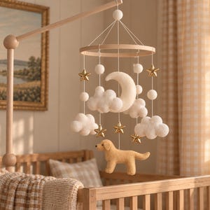 Könnte beinhalten: Ein hängendes Mobile mit einem hellen Holzrahmen, an dem weiße Filzwolken, ein weißer Filzmond, goldfarbene Sterne und eine beige Filzhundefigur an weißen Schnüren von einem hölzernen Arm über einem Kinderbett hängen.