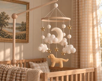 Könnte beinhalten: Ein hängendes Baby-Mobile mit einem hellen Holzrahmen, weißen Filzwolken, einem zentralen weißen Filzmond, goldfarbenen Sternen und einer beigen Filzhundefigur, die an weißen Schnüren an einem Holzarm über einem Kinderbett befestigt ist.